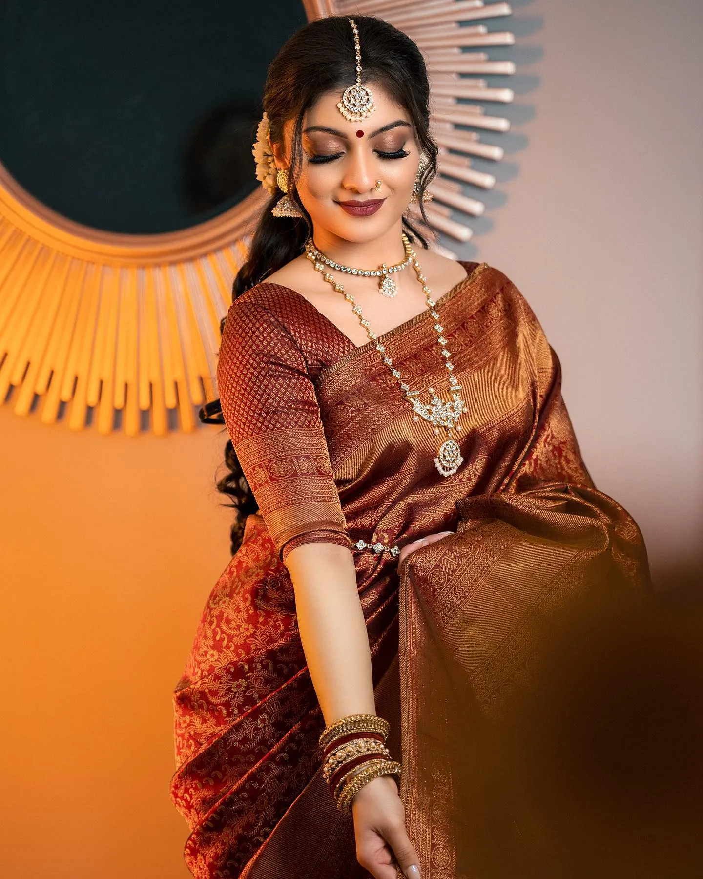 Bride in a maroon saree with gold border, stone necklaces, maang tikka and red and gold bangles