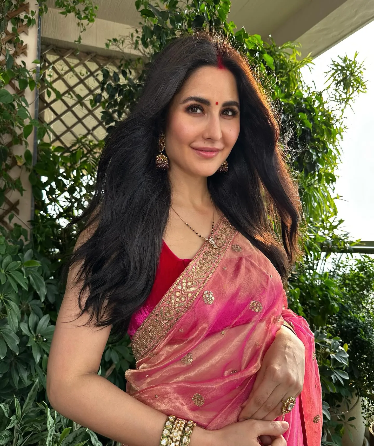 Woman in a pink sheer saree with gold border, jhumkas, mangalsutra and sindoor, smiling outdoors